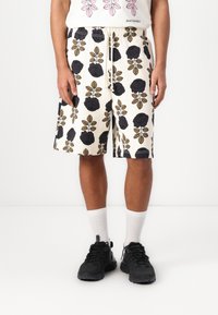 Hvide shorts med et blomsterdesign, der har mørke roser og blade i sort og gyldne nuancer, parret med hvide ankelsokker og sorte sneakers.