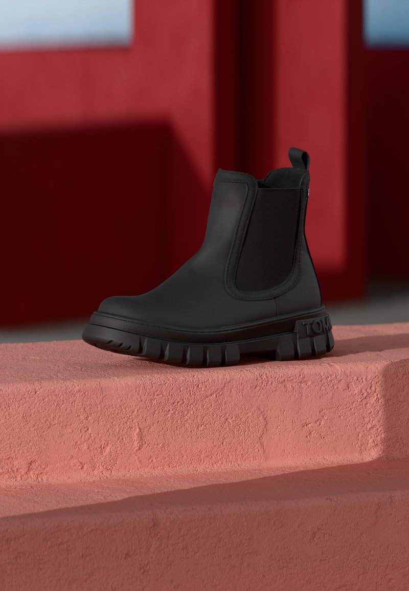 Cizmă Chelsea neagră, realizată dintr-un material neted, având panouri elastice pe laterale și o talpă robustă cu striații pronunțate și detalii de brand.