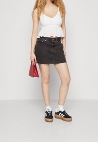 Žena v bílém tílku bez rukávů a černé denimové mini sukni, držící malou červenou kabelku, stojící v černobílých teniskách.