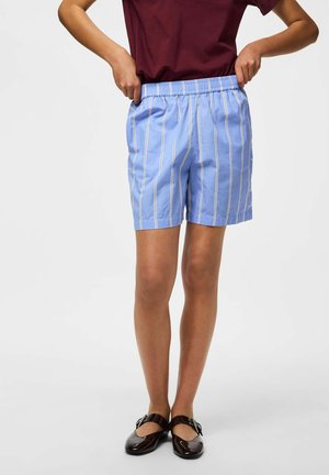 Personne ajustant un short rayé bleu clair, portant un haut bordeaux et des chaussures Mary Jane marron foncé sur un fond uni.
