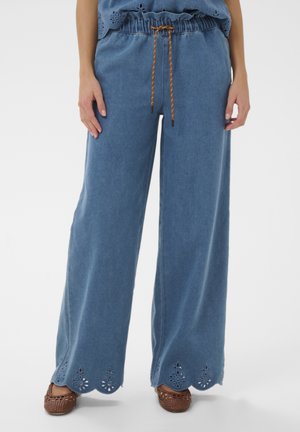 Wijde blauwe broek met geschulpte, geborduurde zoom, bruine taille met trekkoord, gedragen met bruine gevlochten schoenen door een staand persoon.