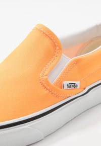 Chaussure orange en toile modèle slip-on avec accents blancs, semelle noire, et détails de couture. Comprend une petite étiquette de marque "VANS".