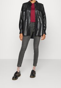 Persona vestida con chaqueta de camisa de cuero sintético negro, top rojo, pantalones ajustados gris de tiro alto, calcetas transparentes con lunares y zapatos negros con hebilla.