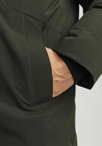 Chaqueta acolchada de color verde oscuro con un gran bolsillo y un puño acanalado. La tela parece suave con detalles de costura visibles.