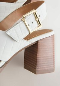 Sandale à talon en cuir blanc avec texture crocodile, bride à boucle dorée, semelle intérieure beige et talon en bois empilé épais.