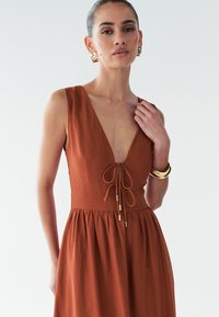 Sukňa bez rukávov v rust-oranžovej farbe s hlbokým výstrihom do V, nastaviteľným viazaním vpredu a rozšírenou sukňou. Hladká tkanina. Zlaté kovové prvky.