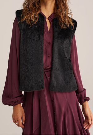 Frau trägt eine ärmellose schwarze Kunstpelzweste über einem langärmeligen, bordeauxfarbenen Kleid mit Knöpfen, plissiertem Rock und gesammelten Manschetten.