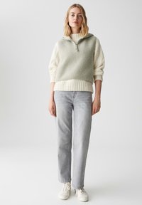 Hellgrüner Fleece-Pullover mit halbem Reißverschluss und geripptem Saum, über einem weißen Pullover getragen, kombiniert mit hellgrauen Jeans und weißen Turnschuhen.