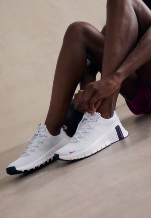 Persona sentada en el suelo atándose unos zapatos deportivos blancos con suelas negras y logotipos rosas de Nike.