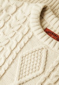 Ecrufarbenes Strickpullover mit Zopfmuster und Diamantdesign. Rippkragen und ein sichtbares Markenetikett mit "Superdry Co".