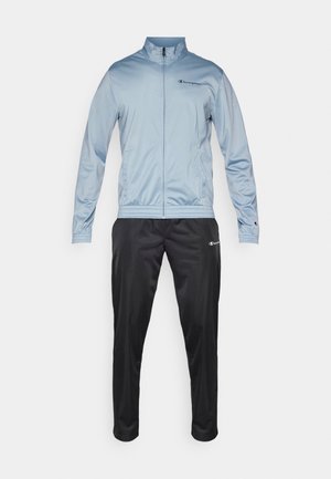 Hellblaues Trainingsjackett mit Reißverschluss, Stehkragen und schwarzem Logo; kombiniert mit schwarzen Trainingshosen mit einem Logo auf dem linken Oberschenkel.