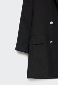 Manica di blazer nero e parte anteriore inferiore con due bottoni tondi argentati e un flap tasca su uno sfondo chiaro.
