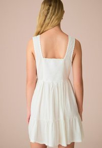 Hvid sommerkjole med en tætsiddende top og en flagrende nederdel, der har scallop-formede stropper og et struktureret stof. Designet omfatter en lagdelt kant.