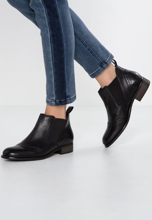 Personne portant un jean bleu foncé et des bottines en cuir noir à talons bas en bois, debout sur un sol blanc.