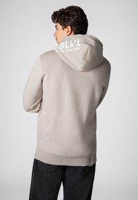 Licht beige hoodie met een gestructureerde afwerking, voorzien van een grote capuchon en witte branding op de achterkant. Gepaard met donkere broeken en een subtiel logo.