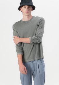 Chemise grise à manches longues en tissu doux, avec un col rond et des poignets retroussés, assortie à un pantalon plissé bleu clair.