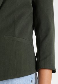 Blazer vert olive au design ajusté. Doté d'une poche unique, de manches retroussées et d'un tissu lisse et texturé. Style décontracté à associer avec un jean.