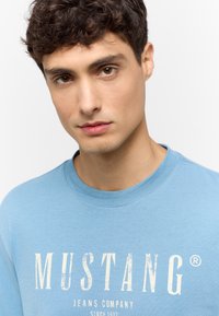Light blue cotton T-shirt featuring a round neckline and distressed white text reading "MUSTANG JEANS COMPANY" across the front.