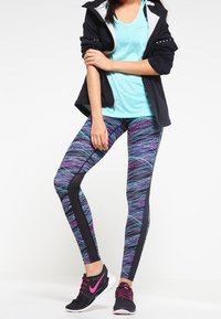 Tenue de sport comprenant une veste noire, un débardeur aqua et un legging à motifs multicolores avec des accents noirs. Des baskets noires complètent le look.