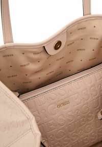 Borsa a tracolla rosa Tetonic con una superficie testurizzata, impreziosita da dettagli dorati. L'interno presenta una fodera beige con un motivo ripetuto "GUESS".