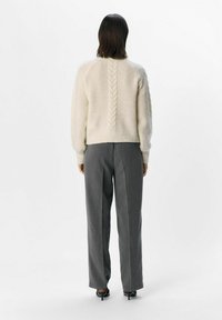 Elfenbensfarvet strikket sweater med tekstureret kabeldetalje ned ad midten og ribbede manchetter, båret med grå bukser og hæle. Bagfra.