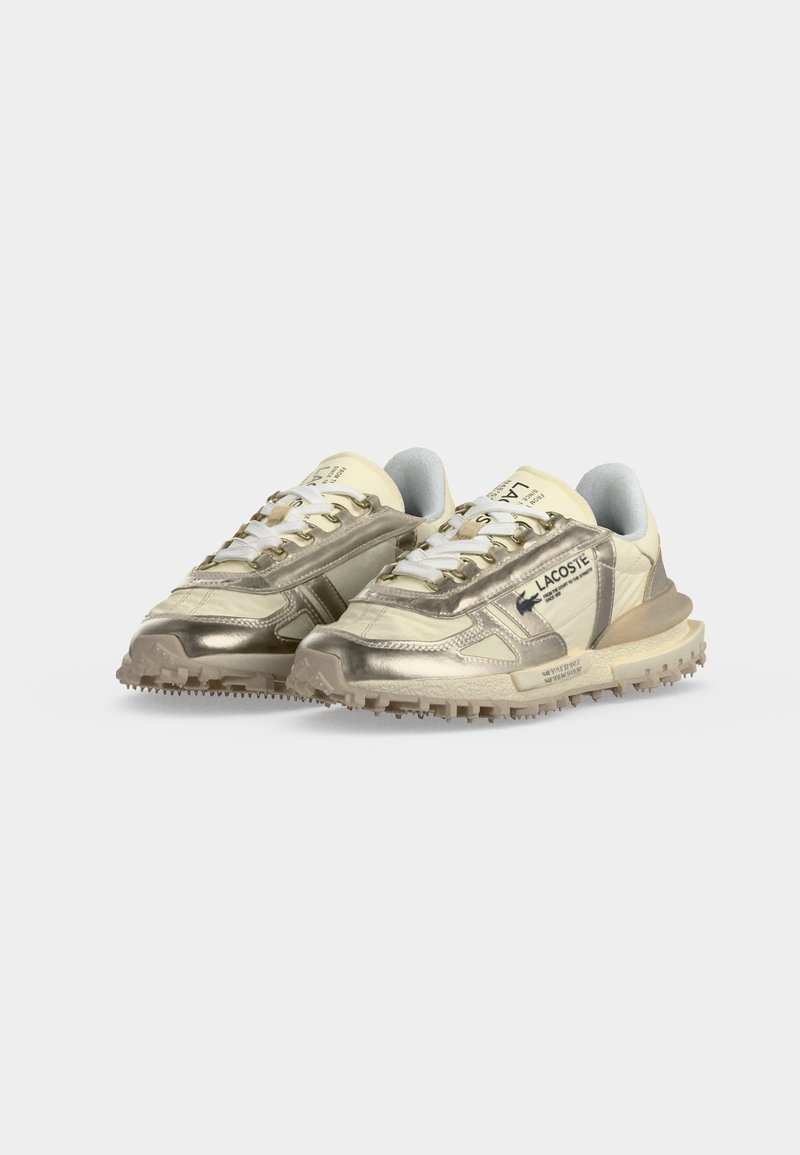 Ein Paar beige und metallisch goldene Lacoste-Sneakers mit weißen Schnürsenkeln und robusten beigen Sohlen, nebeneinander auf grauem Hintergrund gezeigt.