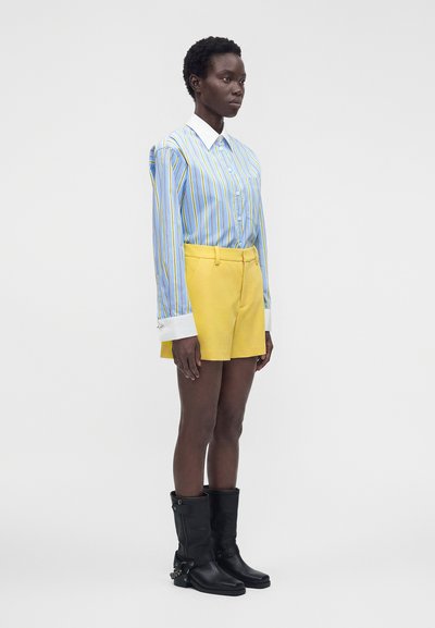 Personne portant une chemise rayée bleue et jaune, un short jaune et des bottes noires mi-mollet, debout devant un fond clair uni.