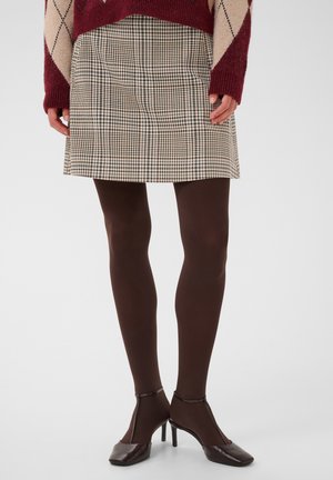 Personne portant une jupe à carreaux beige, un pull bordeaux et beige, des collants opaques marron et des chaussures à talons marron, debout devant un fond uni.