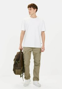 Witte T-shirt met korte mouwen gecombineerd met olijfgroene cargobroek en een bruine rugzak. Witte sneakers maken de outfit compleet. Eenvoudig en functioneel ontwerp.