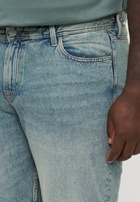 Jeans in denim azzurro chiaro presentano un classico design a cinque tasche, dettagli cuciti e una leggera sfumatura. Il tessuto mostra una consistenza morbida.