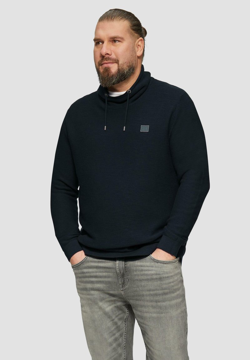 Marinefarbener Pullover mit Kapuze, ausgestattet mit Kordeln, gerippten Bündchen und einem kleinen Logopatch auf der Brust; kombiniert mit hellgrauen Jeans.