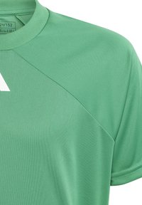 Camiseta deportiva corta verde de manga corta con cuello redondo y detalles de costura visibles cerca del hombro, mostrando parcialmente un logo blanco y la etiqueta de tamaño en el interior del cuello.