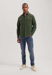 Green button-up shirt with two chest pockets, paired with blue jeans and beige sneakers. The shirt features a textured finish and a collared neck.