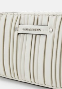 Gros plan d'un sac à main en cuir blanc plissé avec des clous argentés et une plaque métallique portant le logo KARL LAGERFELD.