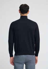 Maglione a collo alto in tessuto a maglia nera, con maniche lunghe e polsini a costine, abbinato a pantaloni grigi chiari. Texture liscia.