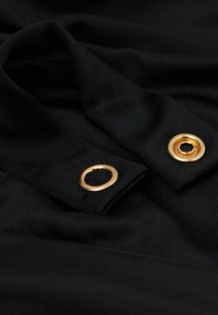 Tela negra acanalada con dos ojales metálicos dorados cerca de los bordes doblados, mostrando textura y detalles metálicos brillantes.