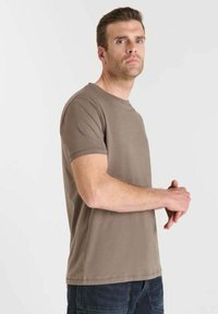 Uomo con capelli castani corti che indossa una semplice maglietta beige a maniche corte e pantaloni scuri, in piedi con le mani unite su uno sfondo bianco.