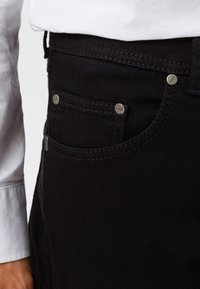 Primer plano de un bolsillo de jeans de mezclilla negra con remaches plateados y parte de la manga de una camisa blanca de manga larga visible.