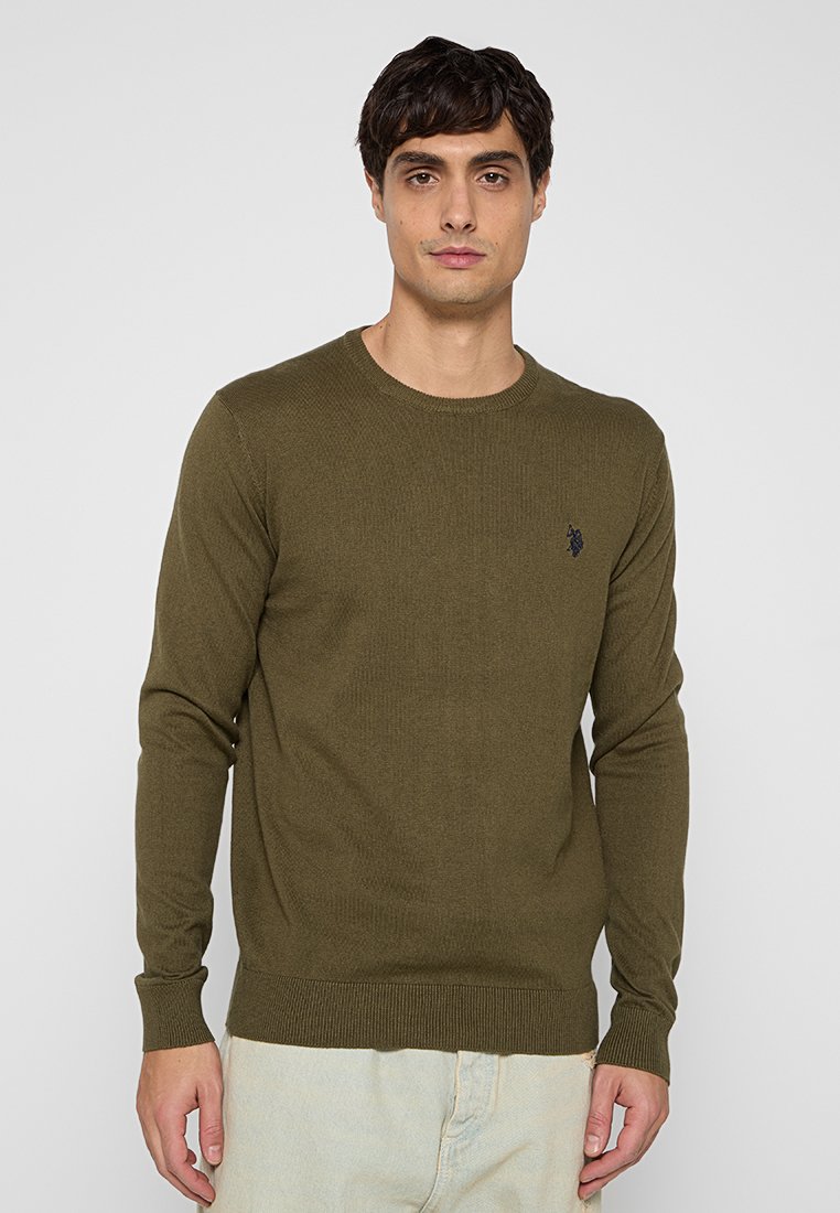 Giovane in maglione a girocollo verde oliva con un piccolo emblema scuro sul petto, in piedi contro uno sfondo chiaro e semplice.
