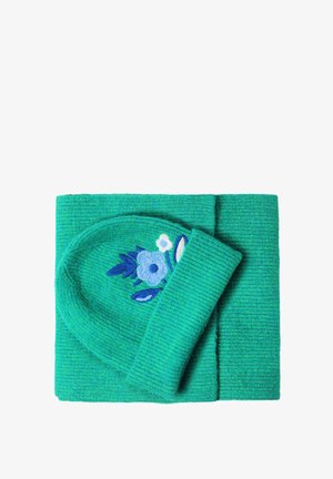 Un bonnet en tricot vert avec des broderies florales bleues, accompagné d'une écharpe pliée assortie sur un fond blanc.