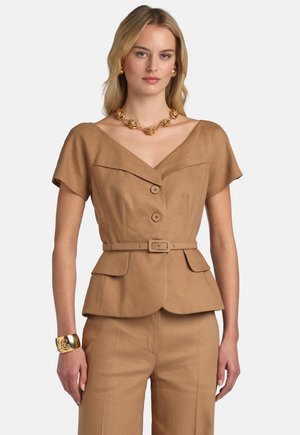 Mujer con blazer marrón claro de manga corta con cinturón y botones, y pantalones a juego, complementada con collar, pendientes y pulsera dorados.