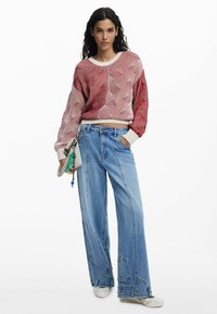 Rot-weißes Wellenmuster-Strickpullover mit weitem Ausschnitt, kombiniert mit hellblauen, weiten Jeans. Eine Hand hält eine kleine graue Tasche und Accessoires.