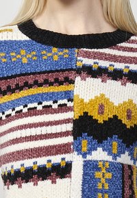 Maglione lavorato a maglia con una serie di motivi colorati, comprese strisce e forme geometriche. Il materiale è strutturato e presenta un collo alto.