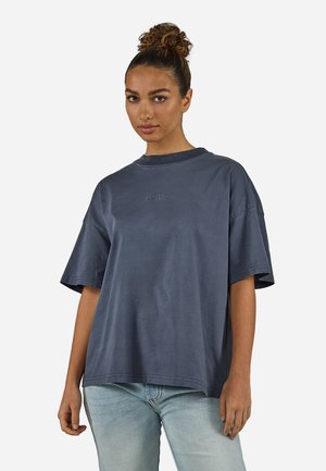 Kvinne med hår i en knute, iført en løs, mørkegrå T-skjorte og lyseblå jeans, med fronten vendt mot en enkel hvit bakgrunn.