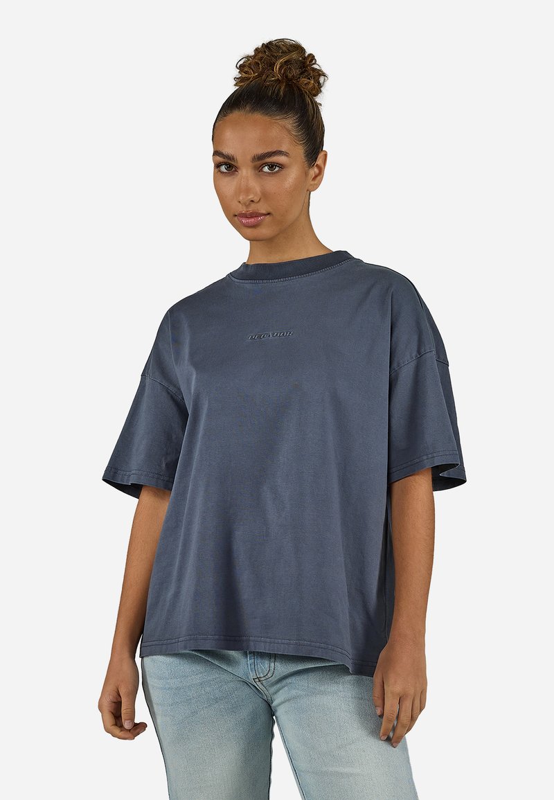 Γυναίκα με τα μαλλιά της δεμένα σε κότσο, φορώντας μια φαρδιά σκούρα γκρι μπλούζα και ανοιχτό μπλε τζιν, που κοιτάζει μπροστά με φόντο έναν απλό λευκό τοίχο.