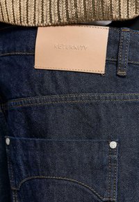 Jeans in denim blu scuro con una targhetta in pelle tan sul giro vita impreziosita dalla scritta "RETERNITY". Presentano dettagli in cucitura dorata e un rivetto argento.