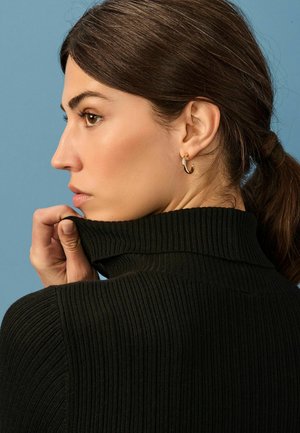 Pull en maille côtelée noir avec un col montant et un tissu texturé. Les cheveux sont attachés, révélant une boucle d'oreille en or.