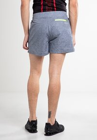 Pánské modré sportovní šortky v melírované textuře, s malou zelenou kapsou na zip vzadu. Kombinované s černými teniskami.