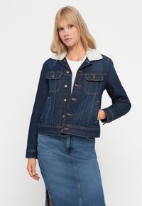 Giacca di jeans di colore blu scuro con colletto in morbido pelliccia bianca. Presenta tasche al davanti, chiusure a bottone e una vestibilità aderente. Indossata con una gonna di jeans.