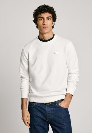 Sudadera blanca de algodón con puños y dobladillo acanalados, que presenta un pequeño logotipo azul en el pecho, usado sobre una camisa navy y jeans azules.
